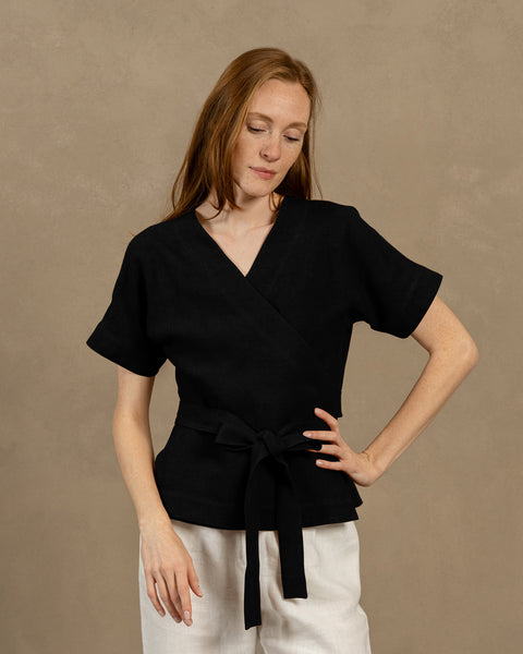 Una persona de piel clara con cabello largo y castaño rojizo, vistiendo una blusa cruzada de lino negra con escote en V, mangas cortas y anchas, y pantalones de color claro. Sostiene los extremos del lazo de la blusa frente a ella, formando un moño.