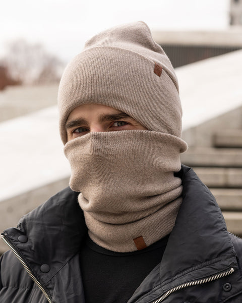 Un modelo masculino, visto de perfil, lleva un gorro de punto color beige cremoso y un braga de cuello a juego que le cubre la nariz y la boca. Tanto el gorro como la braga tienen un pequeño parche de cuero marrón con el logo de Menique. Tiene barba corta y cabello oscuro. El fondo es blanco sólido.