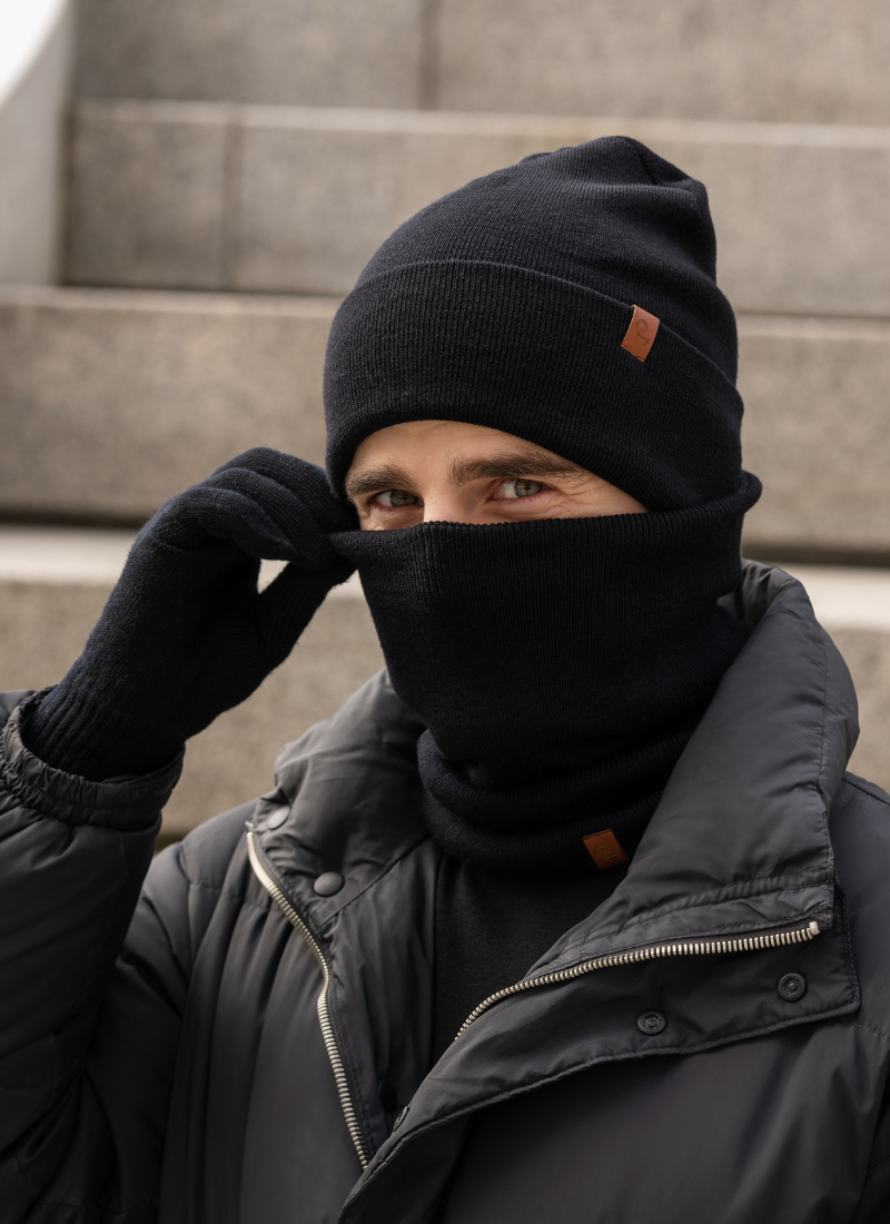A person dressed in all-black winter accessories, including a knit beanie, neck gaiter, and gloves, poses in front of blurred stone steps while wearing a dark jacket.