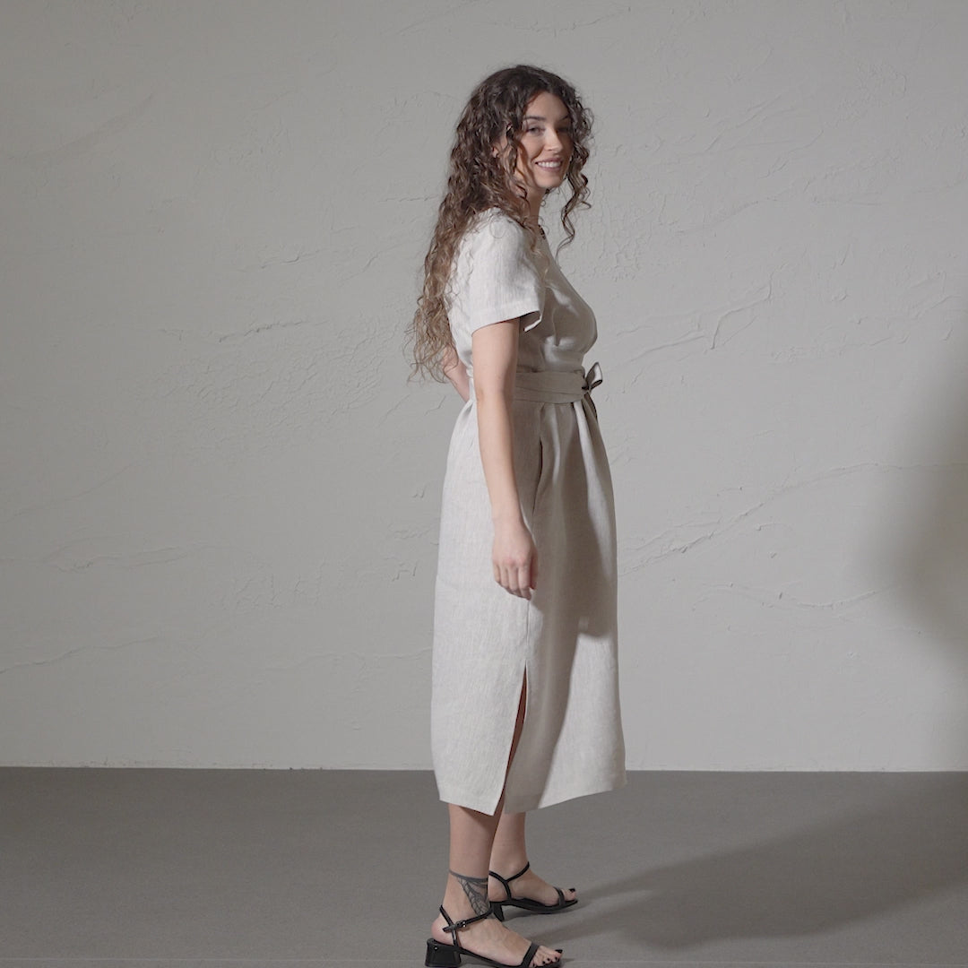 A woman with curly brown hair smiles and models a short-sleeved, natural linen dress with a tie waist and pockets against a textured gray backdrop in a short video.