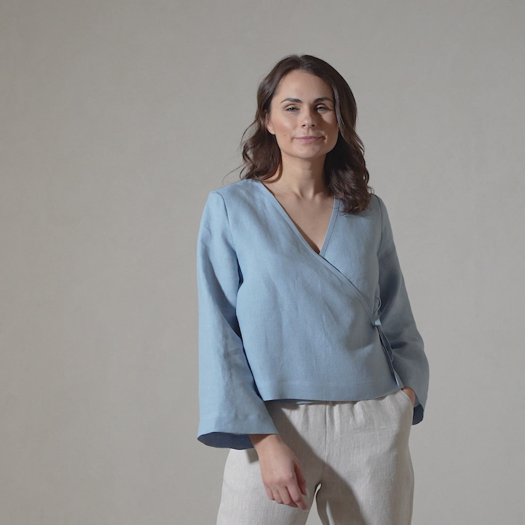 A woman with shoulder-length brown hair models a cloudy blue linen wrap top with long sleeves and linen menique pants, subtly shifting her pose and smiling gently in a short video against a plain background.