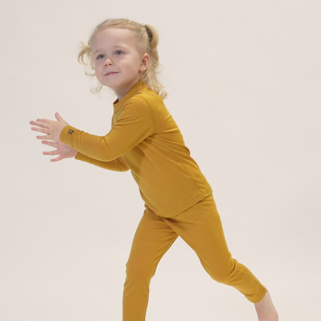 A child with curly blonde hair styled in pigtails, wearing a yellow long-sleeved top and matching pants, is captured mid-movement against a white background. The child is looking up and to the right with a slight smile, and their hands are raised in front of them as if preparing to catch something.