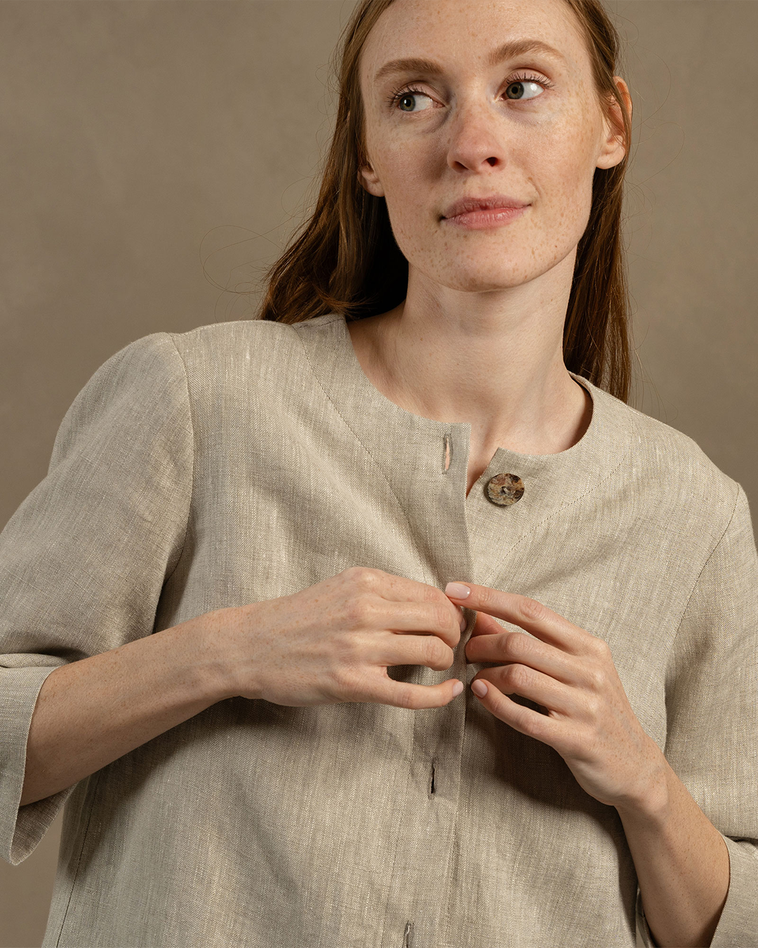 A portrait of a woman with long reddish hair wearing a neutral beige linen shirt, looking softly away from the camera against a grey background.