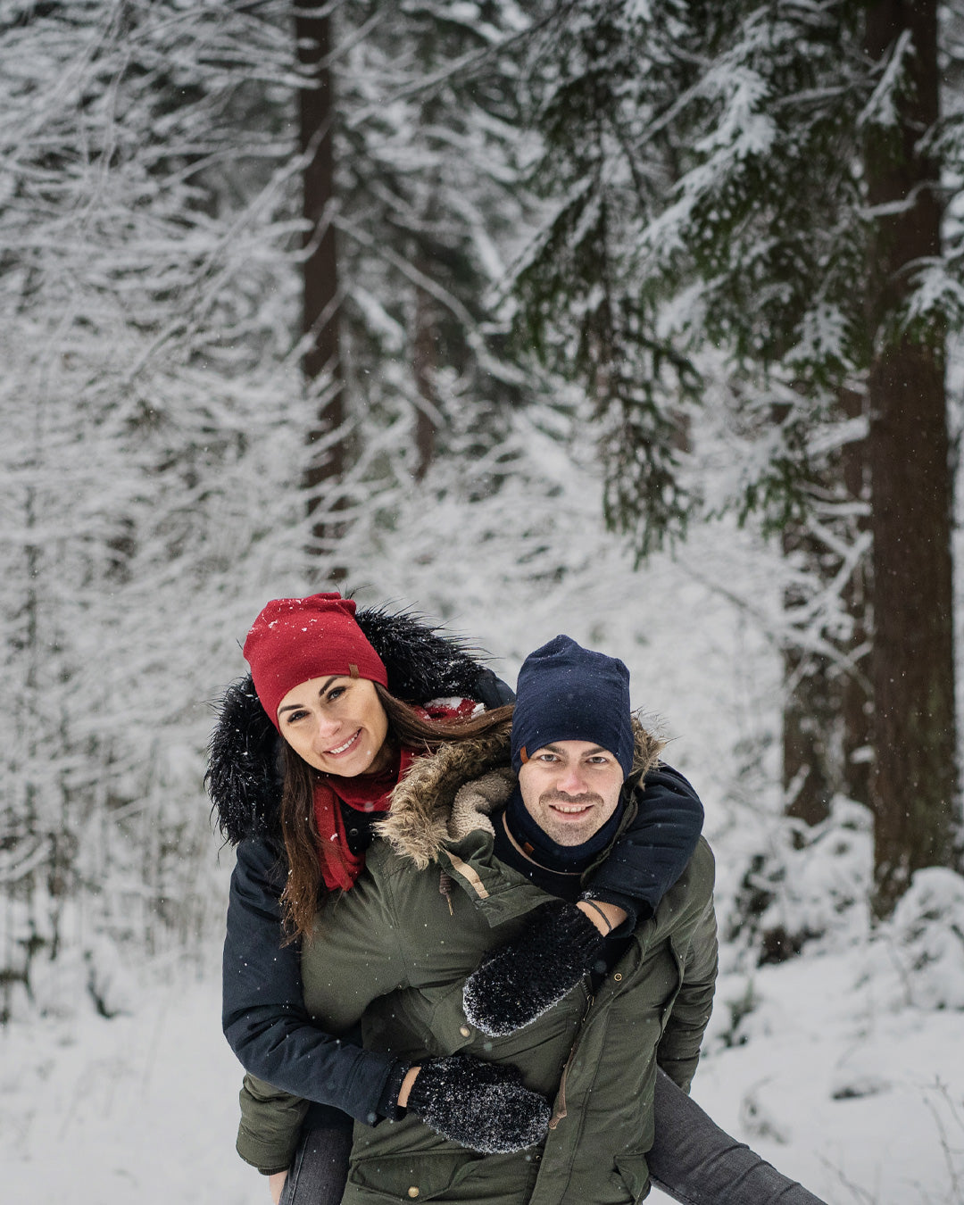 Pareja disfrutando del invierno al aire libre, mujer con gorro rojo abrazando a un hombre por detrás en un bosque nevado