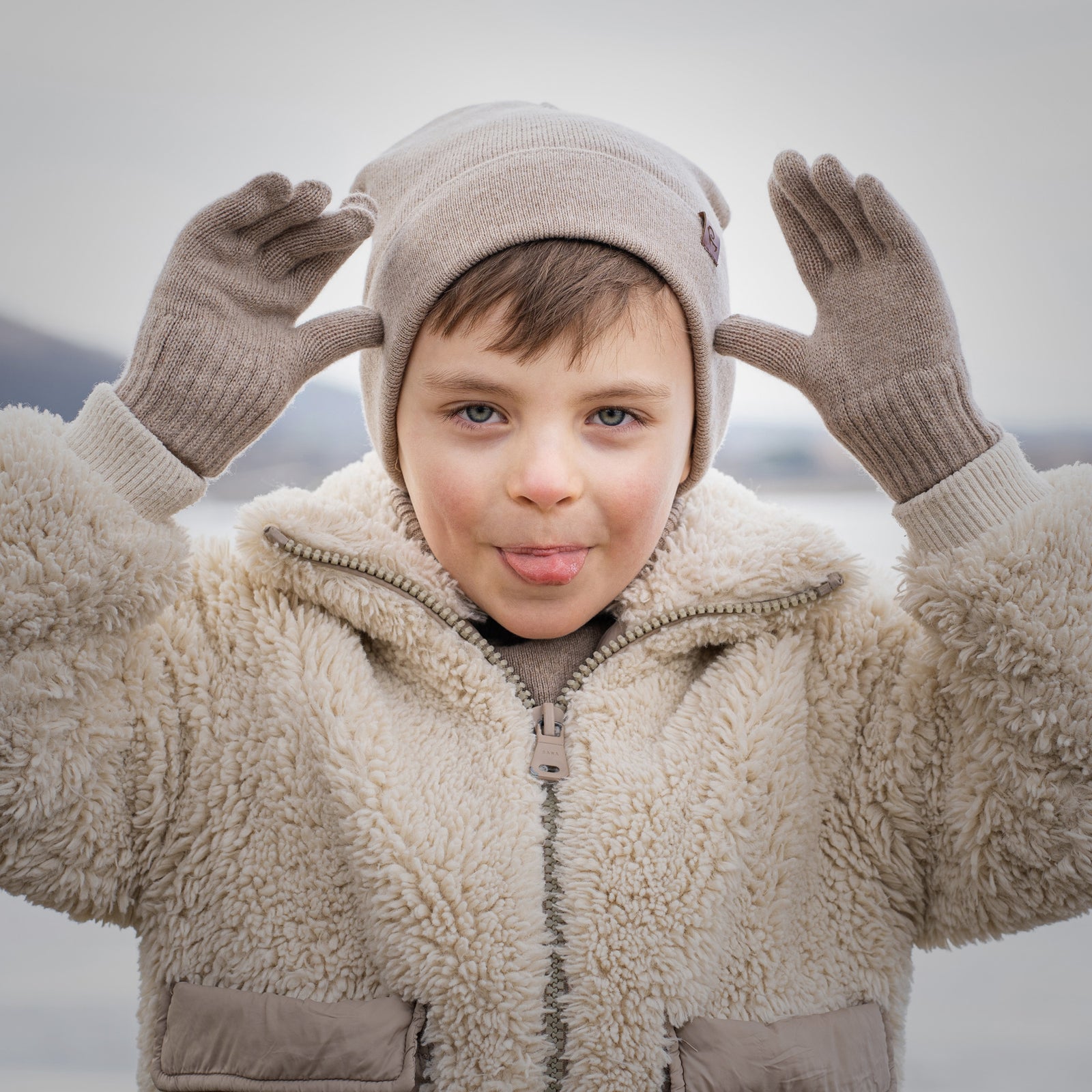 Niño con chaqueta sherpa beige, gorro de punto y guantes, posando de forma juguetona al aire libre en invierno