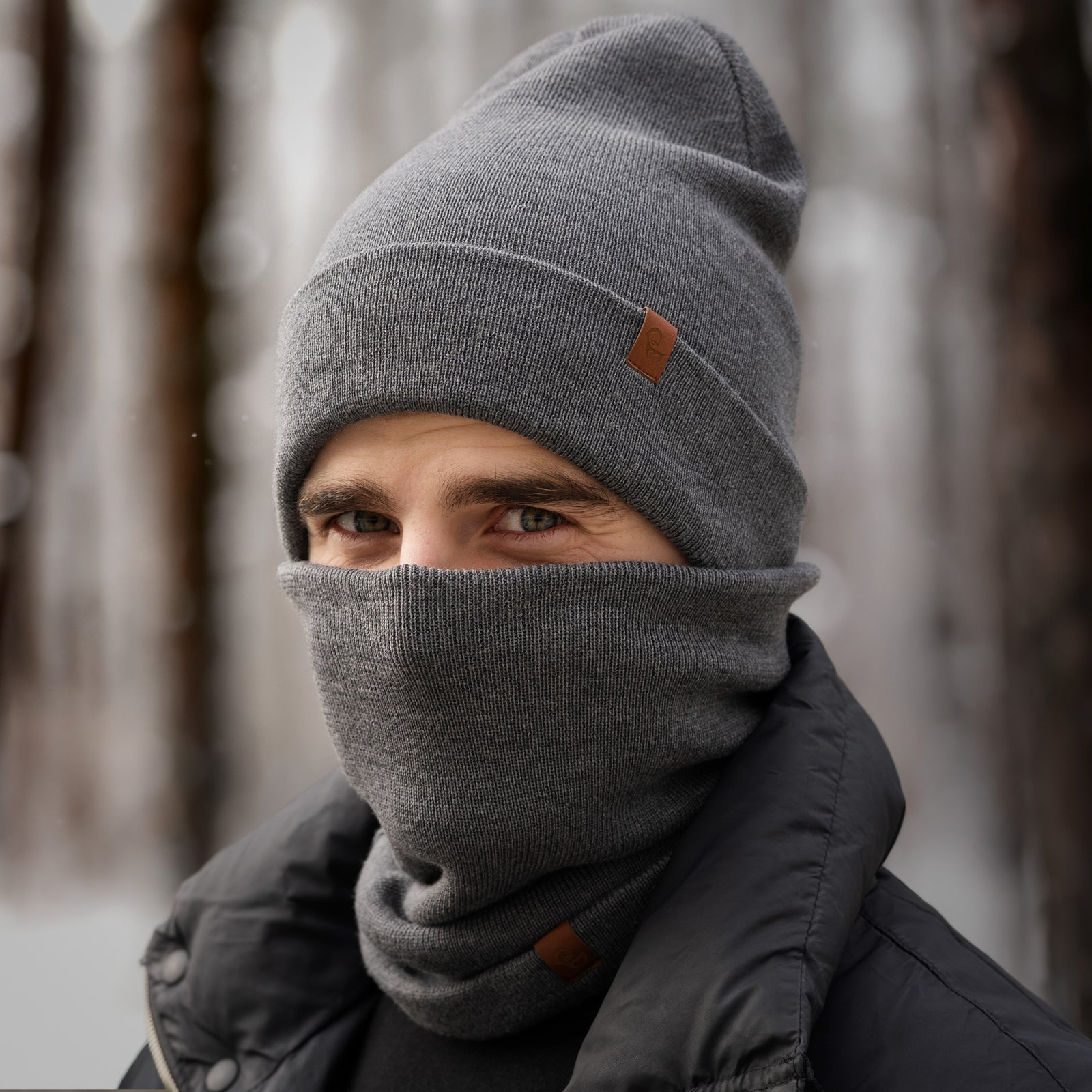 Hombre con gorro de merino gris y braga de cuello, de pie al aire libre en un bosque nevado