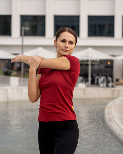 Mujer con una camiseta menique de lana merina en color cereza real estirándose al aire libre con un edificio de fondo