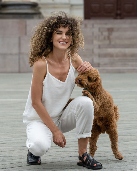 Hermosa mujer posando en la ciudad. Lleva una blusa de lino blanco puro y pantalones de lino blanco puro, acompañada de un perro caniche marrón.