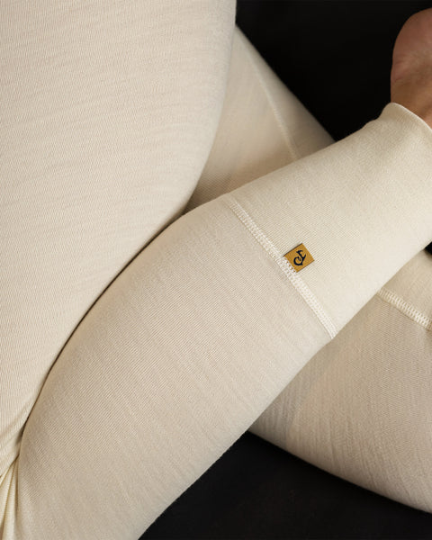 Se muestra un primer plano de un par de mallas de color natural hechas de lana merina. Las mallas tienen una cintura alta y una silueta ajustada. La tela tiene un acabado liso y mate, y está libre de cualquier adorno o patrón visible.