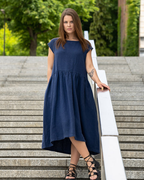 Joven posando en una calle de la ciudad sobre unas escaleras, vistiendo un vestido blusón de lino Cecilia en color azul tormenta. Chica con un tatuaje de gato en su brazo izquierdo.