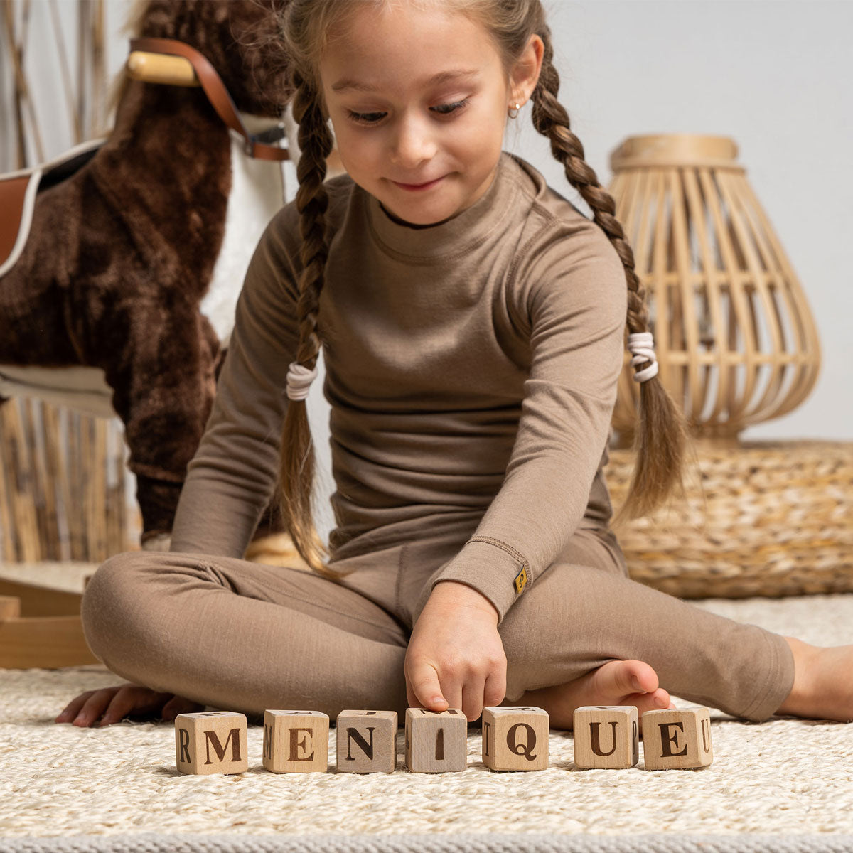 Una niña, sosteniendo un oso de peluche, lleva puestos los pantalones Menique Merino 160 en color beige. Está de pie sobre una cama, sonriendo.